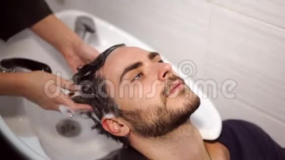 女性应用洗发水按摩头发客户男士在美容院理发师洗头快乐的白种人视频的预览图
