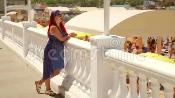 一个穿着蓝色裙子的女人站在长廊上看着海滩和大海视频的预览图