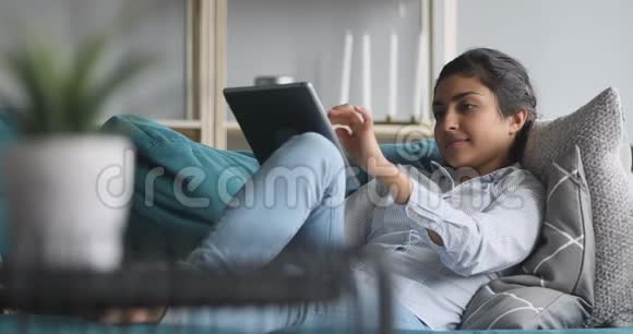 使用数码平板电脑在沙发上放松的印度快乐女人视频的预览图
