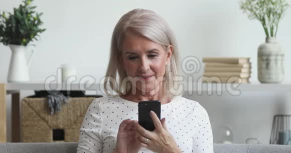 快乐的老女人使用手机手机应用程序坐在沙发上视频的预览图