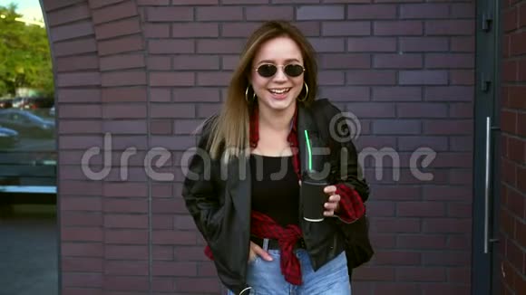 秋天年轻漂亮的女学生站在砖墙附近戴着太阳镜手里拿着咖啡笑着视频的预览图