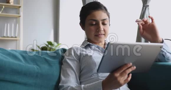 使用数码平板电脑的印度快乐女孩坐在沙发上视频的预览图