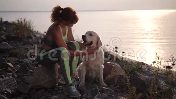 适合的女人抚摸坐在岸边日落的狗视频的预览图