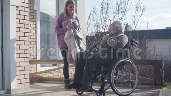 漂亮的高加索女人戴着眼镜为坐在轮椅上的残疾老人带来毯子护士视频的预览图