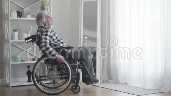 悲伤的老高加索残疾人的肖像从窗户转向轮椅滚动而去抑郁症男性退休人员视频的预览图