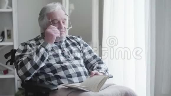 白人老人戴眼镜和看书的肖像坐在轮椅上的残疾退休老人视频的预览图