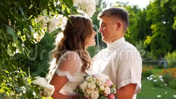 新娘和新郎在茂密的白玫瑰灌木丛附近微笑和亲吻新婚夫妇拥抱在一起彼此相爱视频的预览图