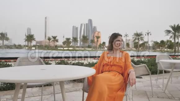 一个身穿橙色连衣裙的女孩坐在一张桌子前背景是阿布扎比的夜晚视频的预览图