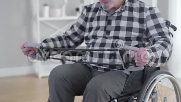 一个不能辨认的白人坐在轮椅上伸展弹力带老年男性残疾人在室内运动视频的预览图