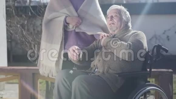 坐在轮椅上的积极的高加索老人在阳光下坐在户外与面目全非的护士交谈视频的预览图