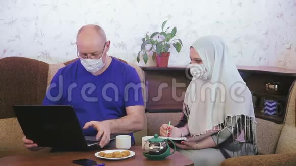 穆斯林夫妇戴着防护面具在家中进行检疫以预防冠状病毒在沙发上从事远程工作视频的预览图