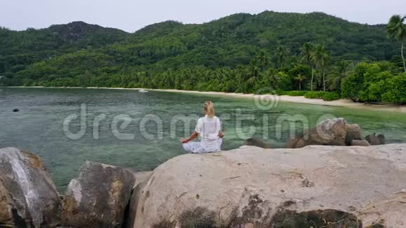 空中飞行围绕金发女孩坐在花岗岩岩石与异国情调的热带海滩和冥想生活方式的平衡视频的预览图