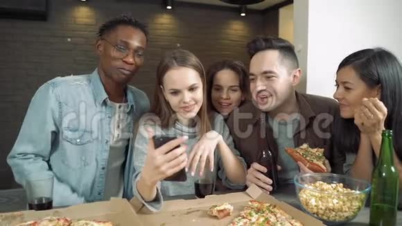 多民族的朋友看着智能手机在酒吧或家里谈笑风生吃披萨和零食视频的预览图