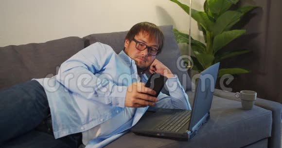 快乐的年轻人躺在家里的沙发上用笔记本电脑和智能手机工作视频的预览图