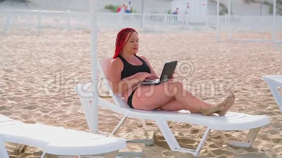 在海滩上一个穿着泳衣留着红色头发的年轻犹太妇女坐在躺椅上手里拿着一台笔记本电脑视频的预览图