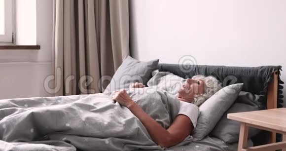 一个人在舒适的床上醒来的年轻快乐的老太太视频的预览图