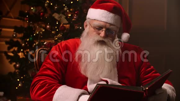 戴着眼镜留着白胡子的圣诞老人在圣诞树和壁炉的背景上读着一本红色的书视频的预览图
