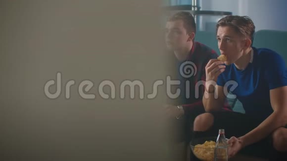 三个穿着蓝色T恤的好朋友坐在沙发上喝啤酒看电视时跳着体育节目视频的预览图