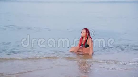一个穿着红色辫子的黑色泳衣的女人坐在海里从迎面而来的海浪中坠落视频的预览图