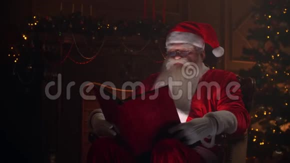 穿着圣诞老人服装的老人坐在大气的房间里读着一本红色封面的神奇闪亮的书视频的预览图