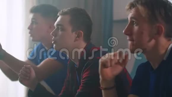 三个穿蓝色T恤的男人和朋友一起坐在沙发上看足球或篮球比赛欢呼和鼓掌视频的预览图