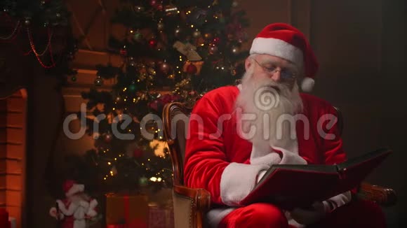 快乐的圣诞老人条款坐在装饰的房间里的摇椅里读着一本红色封面的书节日的心情圣诞节视频的预览图