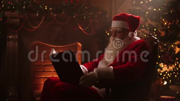 现实的圣诞老人条款坐在壁炉和圣诞树附近打开一本大的魔法书里面有闪亮的书页视频的预览图