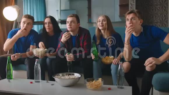 5个穿着蓝色t恤的男人和女人都很担心坐在沙发上看电视吃爆米花视频的预览图