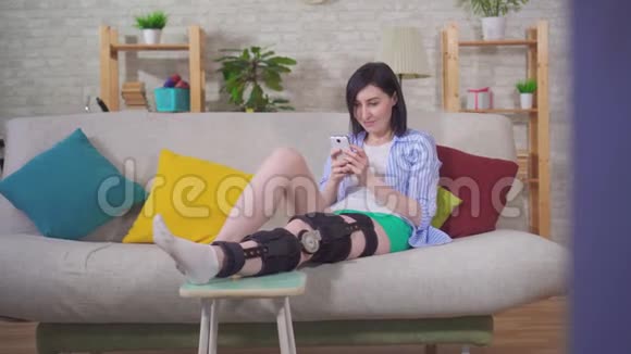 受伤后带手机的女性膝盖弯曲矫形膝盖支架躺在家里的沙发上视频的预览图