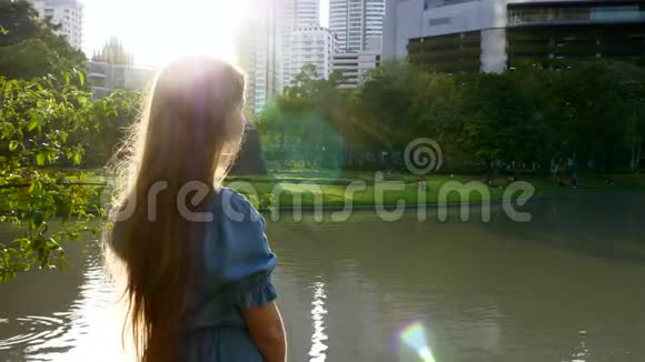 美丽的快乐女孩站在公园的湖边夕阳下的树木视频的预览图