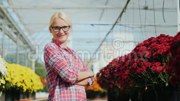 年轻的女人微笑着看着相机它站在植物苗圃里的花的背景上视频的预览图