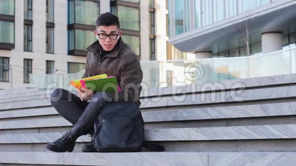 年轻的亚裔男学生坐在大学台阶上看书视频的预览图