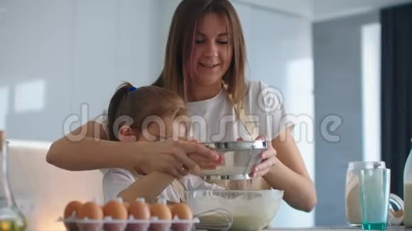 小可爱的女儿正在厨房里帮她的母亲在碗里加入面粉和筛子并混合配料视频的预览图