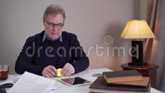 年长的白人男子写笔记并把它们贴在桌子上的物体上戴眼镜的成熟退休人员视频的预览图