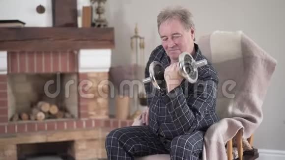 年长的高加索人举着哑铃坐在家里的扶手椅上在职退休人员上午锻炼视频的预览图