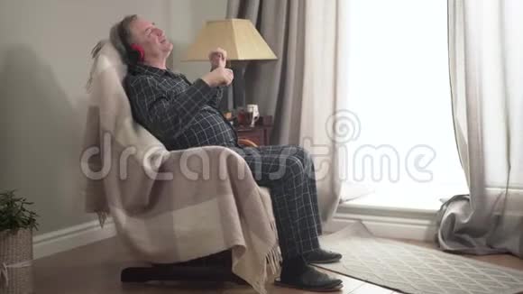 快乐的高白种人坐在扶手椅上戴着耳机在感情上做手势开朗的男性侧观视频的预览图