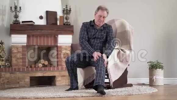年老的白人男子在室内的扶手椅上揉膝老年退休人员健康问题画像老龄化视频的预览图