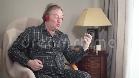 开朗的高白种人穿着睡衣听着耳机里的音乐模仿弹吉他老年人视频的预览图