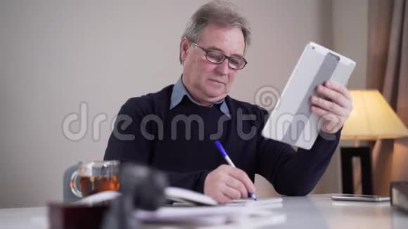 高加索人在接到视频电话时在桌旁写作戴眼镜的成年男性退休者视频的预览图