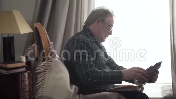 积极成熟的白种人老人坐在窗前的相册和微笑高级退休人员的侧视视频的预览图