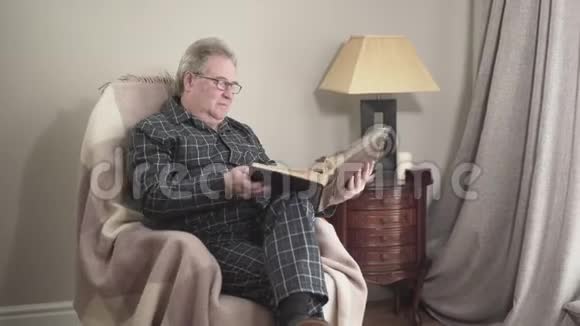 穿着睡衣的老白种人坐在摇椅上看着相册里的照片男性退休人员视频的预览图