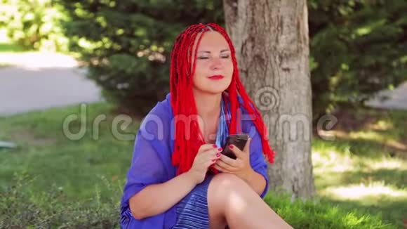 一个年轻的女人坐在公园的草地上在智能手机中的信使中交流视频的预览图
