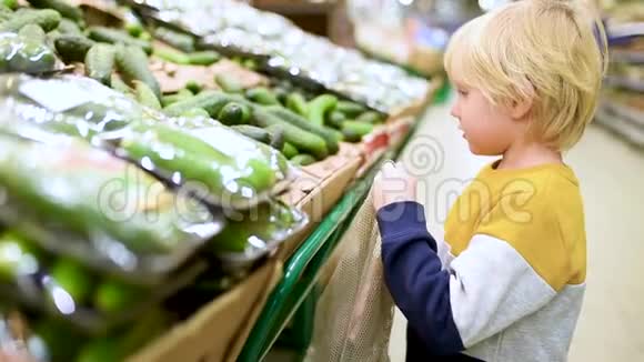 可爱的小男孩选择新鲜的有机黄瓜并放入网状购物袋在食品商店或超市零废物概念视频的预览图