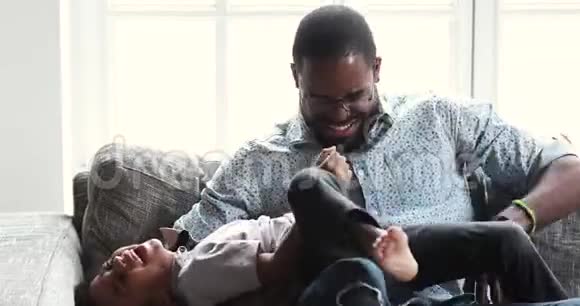 有趣快乐的非洲爸爸在沙发上挠痒可爱的小儿子视频的预览图