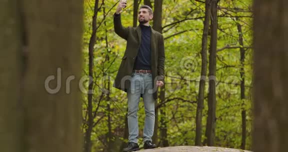 微笑的白种人白发苍苍的男子站在秋天的森林中的树木之间在他的智能手机上拍照视频的预览图