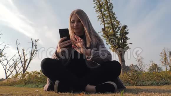 年轻迷人的金发女孩坐在公园的户外用电话视频的预览图