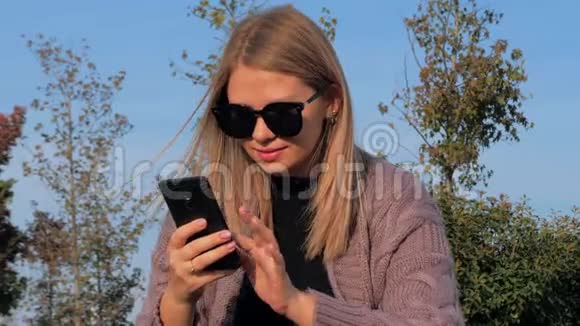 年轻迷人的金发女孩坐在公园的户外用电话视频的预览图