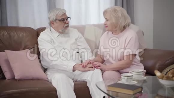 一对年长的白人夫妇坐在沙发上手牵着手说话已婚的成熟配偶在家里聊天视频的预览图