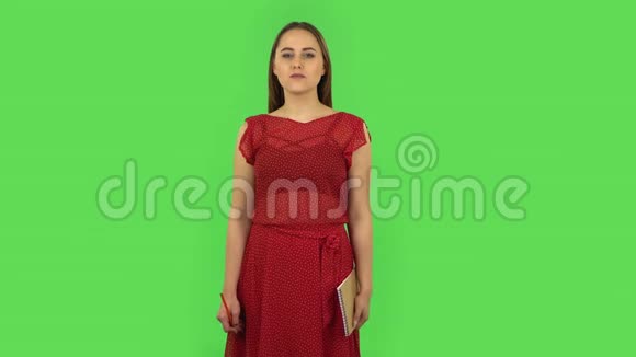 穿着红衣服的小女孩站在那里思考然后用铅笔在笔记本上快乐地写字绿色屏幕视频的预览图