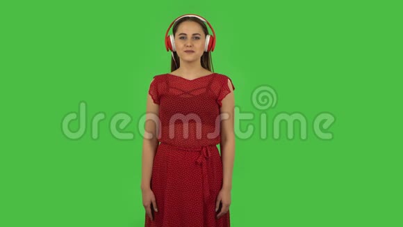 穿着红色裙子的温柔女孩正在用红色的大耳机跳舞和欣赏音乐绿色屏幕视频的预览图